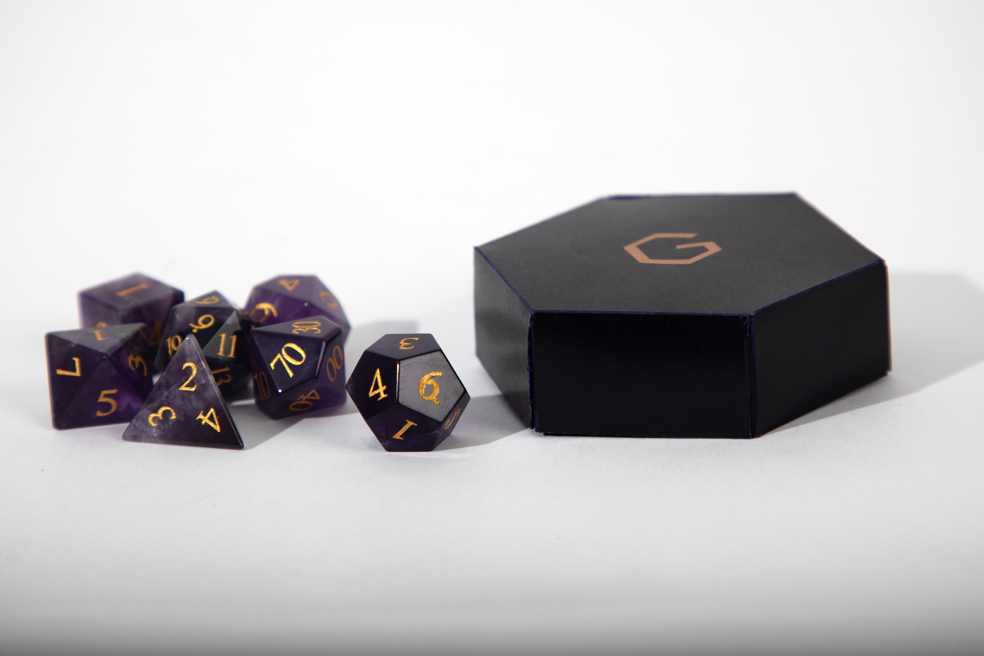 Links ist ein sieben-teiliges Würfelset aus Amethyst mit goldenen Zahlen zu sehen, rechts davon steht eine kleine sechseckige, violette Schachtel mit einem goldenen G am Deckel.